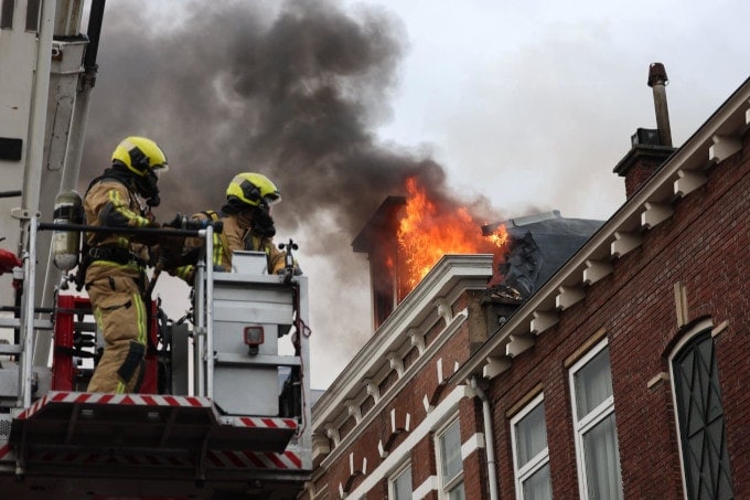 Twee brandweermannen in de bak van een hijskraan met op de achtergrond vlammen die uit het dak van een woning komen.